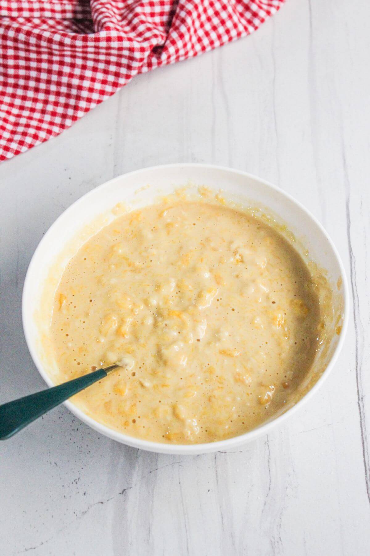 A white bowl filled with a thick, pale yellow batter and a black spoon, placed on a white surface with a red and white checkered cloth nearby.
