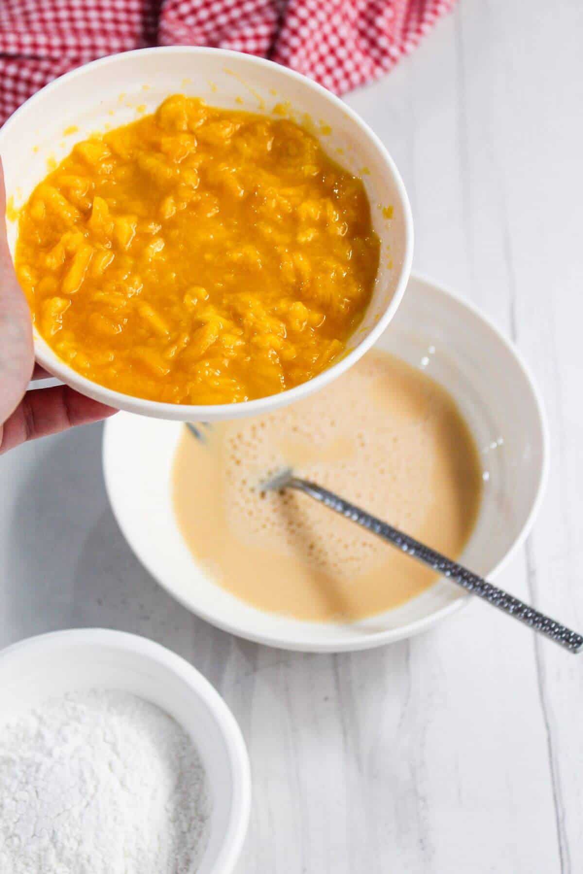 A hand holds a bowl of mashed pumpkin over a mixing bowl with liquid and a whisk; a small bowl of flour sits nearby on a white surface.