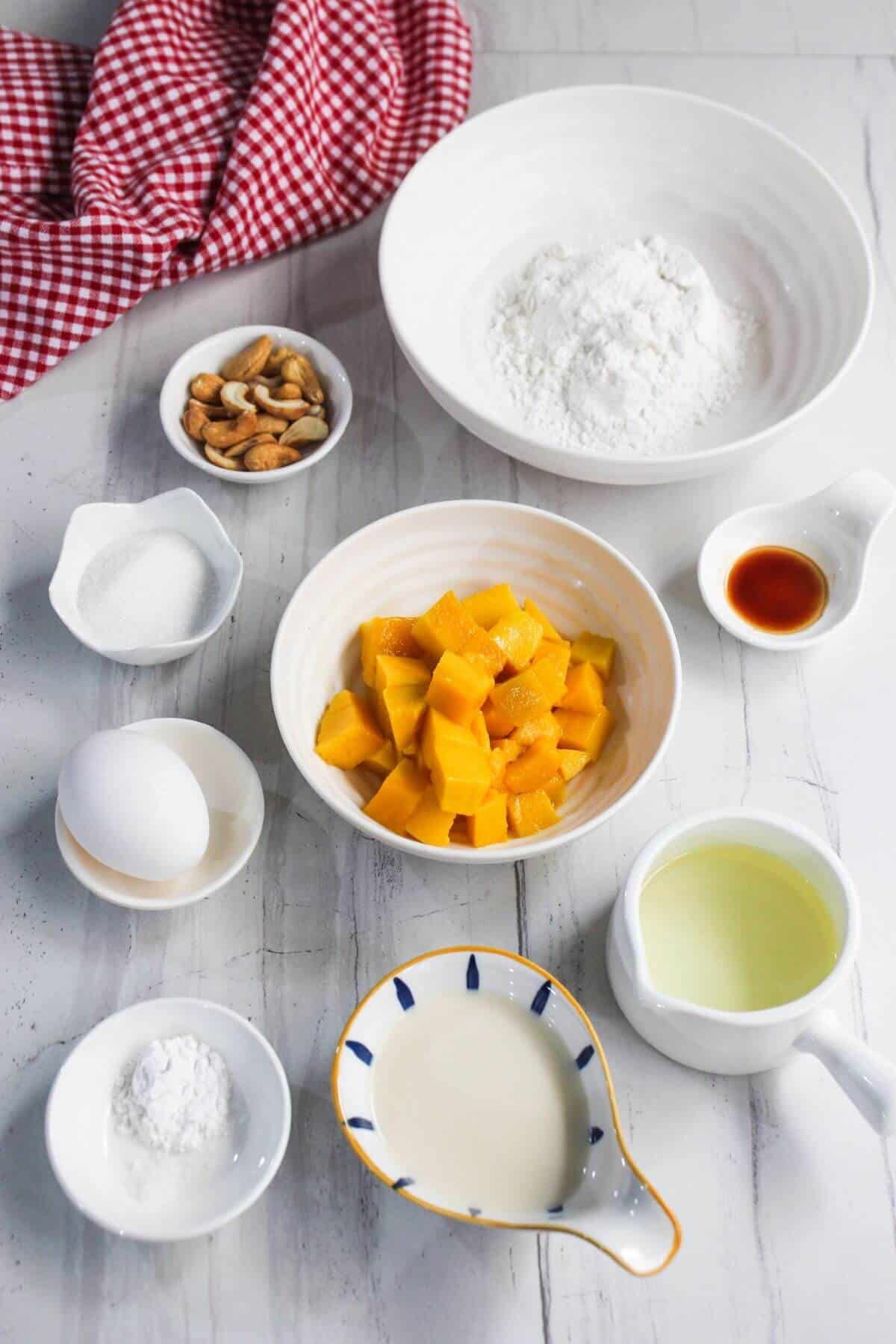 Assorted baking ingredients including flour, cubed mango, sugar, egg, milk, oil, cashews, vanilla extract, and baking powder arranged on a white surface with a red checkered cloth.