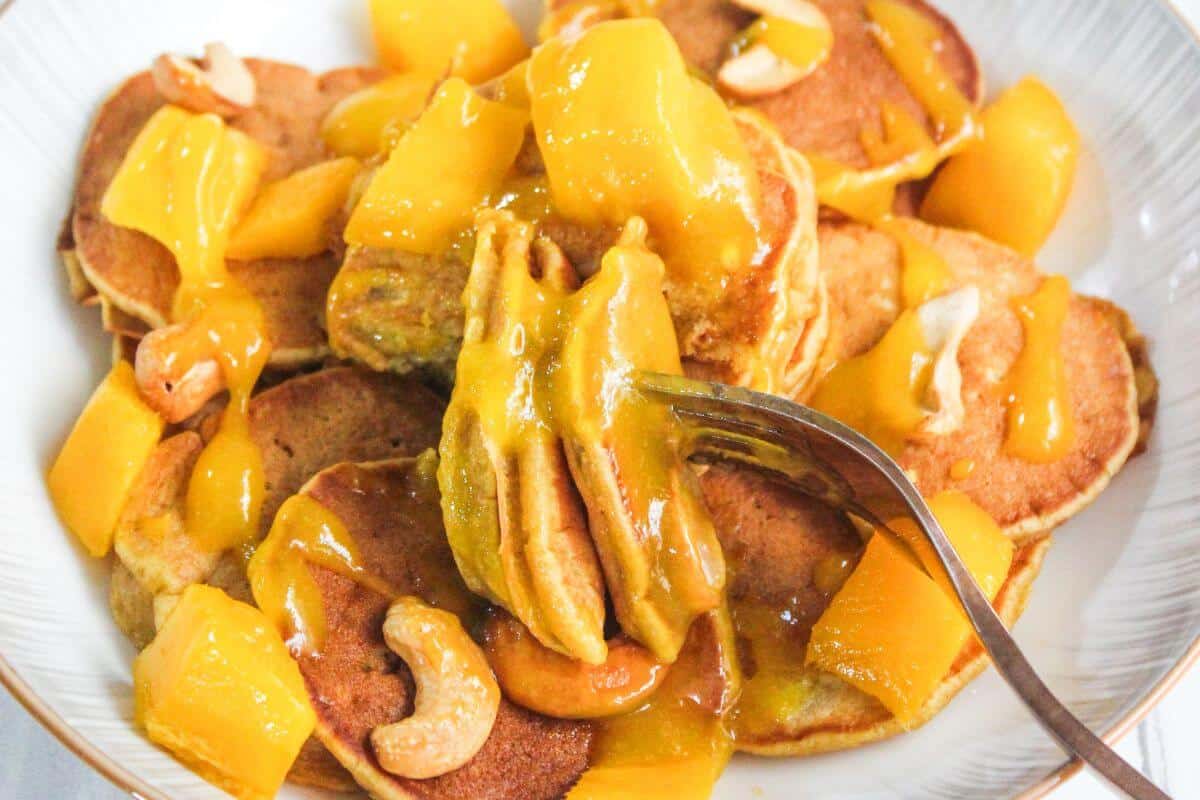 A plate of pancakes topped with mango chunks, cashew nuts, and a glossy mango sauce, with a fork holding a bite-sized piece.
