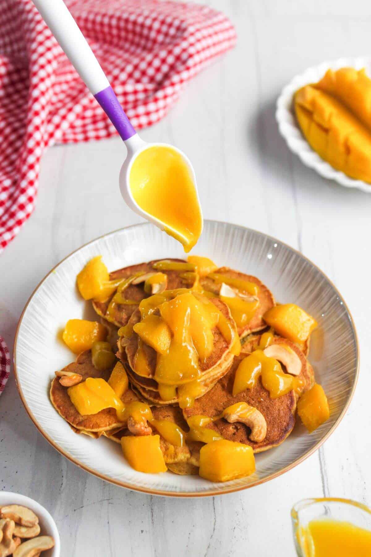 A spoon drizzles mango sauce over a stack of pancakes topped with diced mango and cashews on a white plate, with a red checkered cloth in the background.