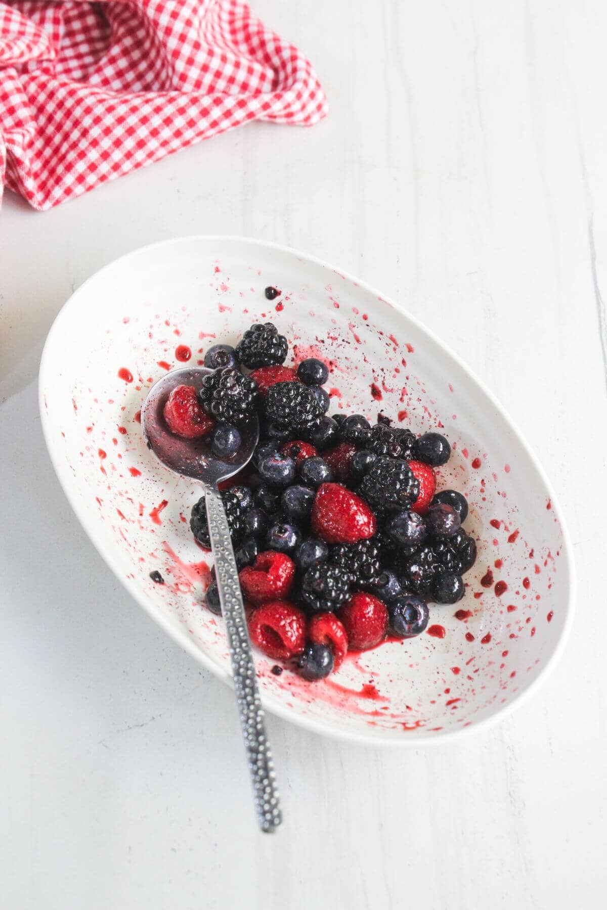 An oval white dish with mixed fresh berries-blackberries, blueberries, raspberries, and strawberries-partially eaten, with a spoon and red juice stains on the dish.