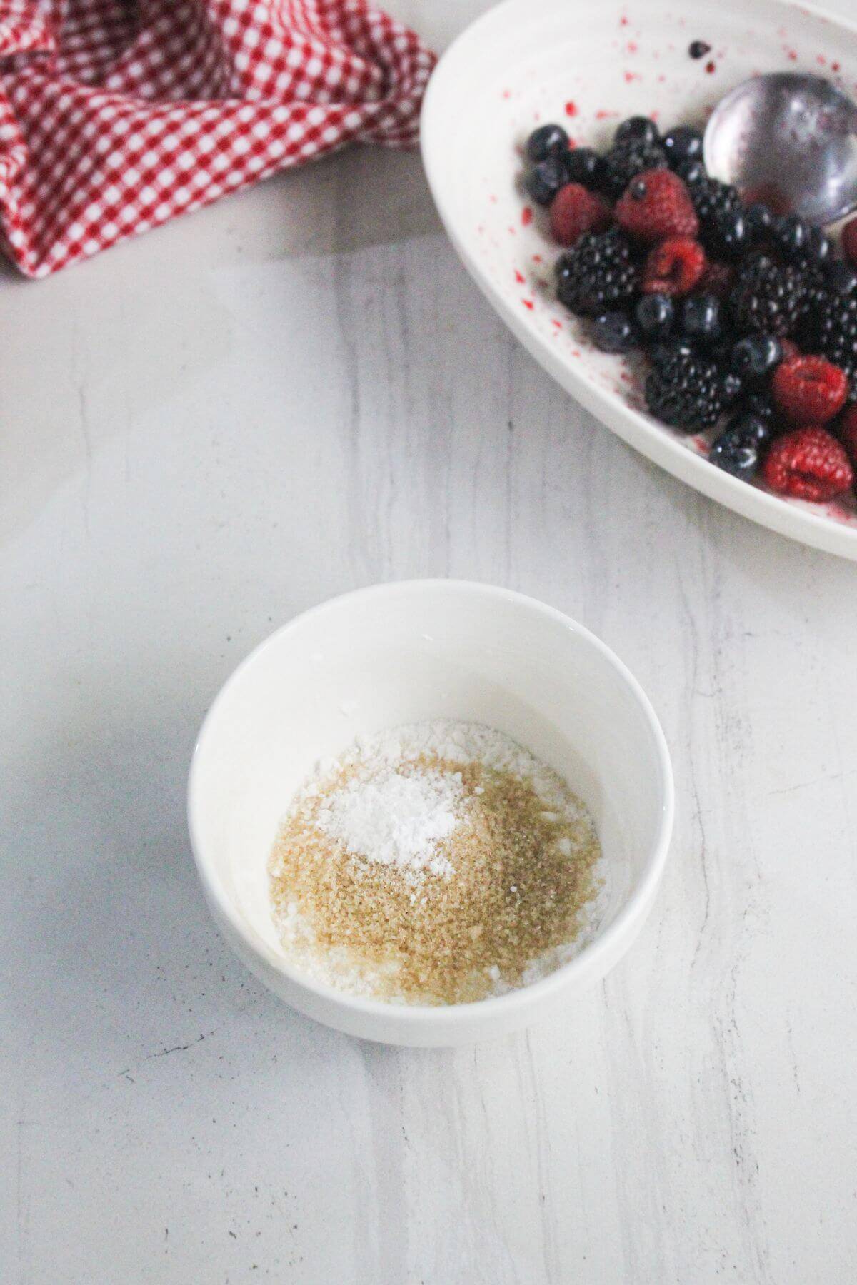 A small white bowl with white and brown sugar sits on a marble surface; a plate of mixed berries and a spoon are in the background.