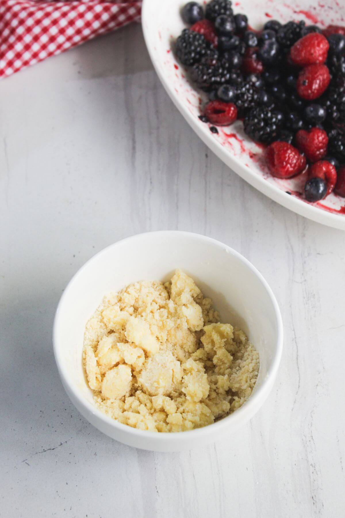 A small white bowl filled with crumbly dough sits on a white surface; in the background, a plate holds a mix of berries and a red checkered cloth.