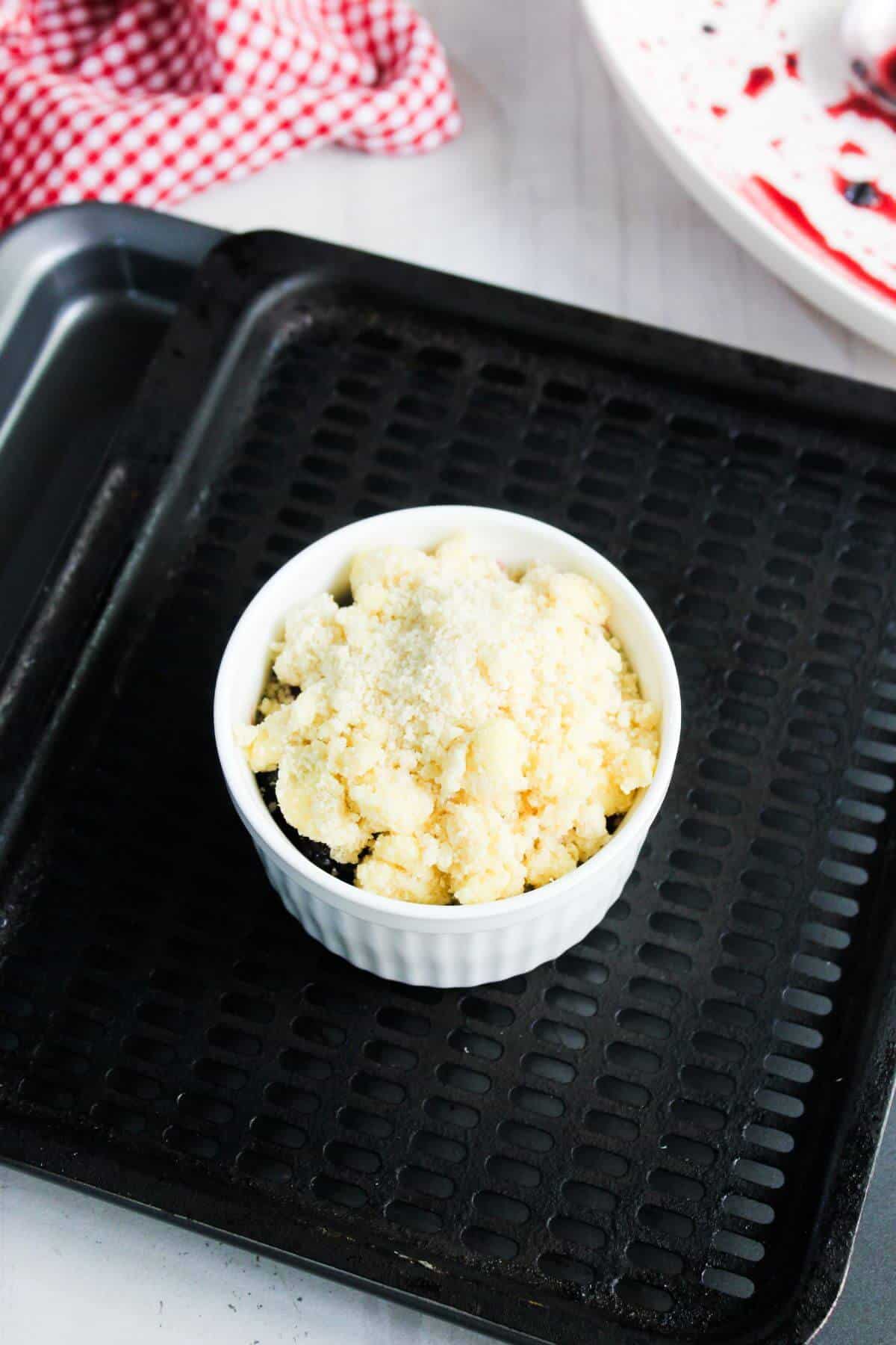 A white ramekin filled with crumbly topping sits on a black baking tray; a red and white cloth and a plate with red stains are in the background.