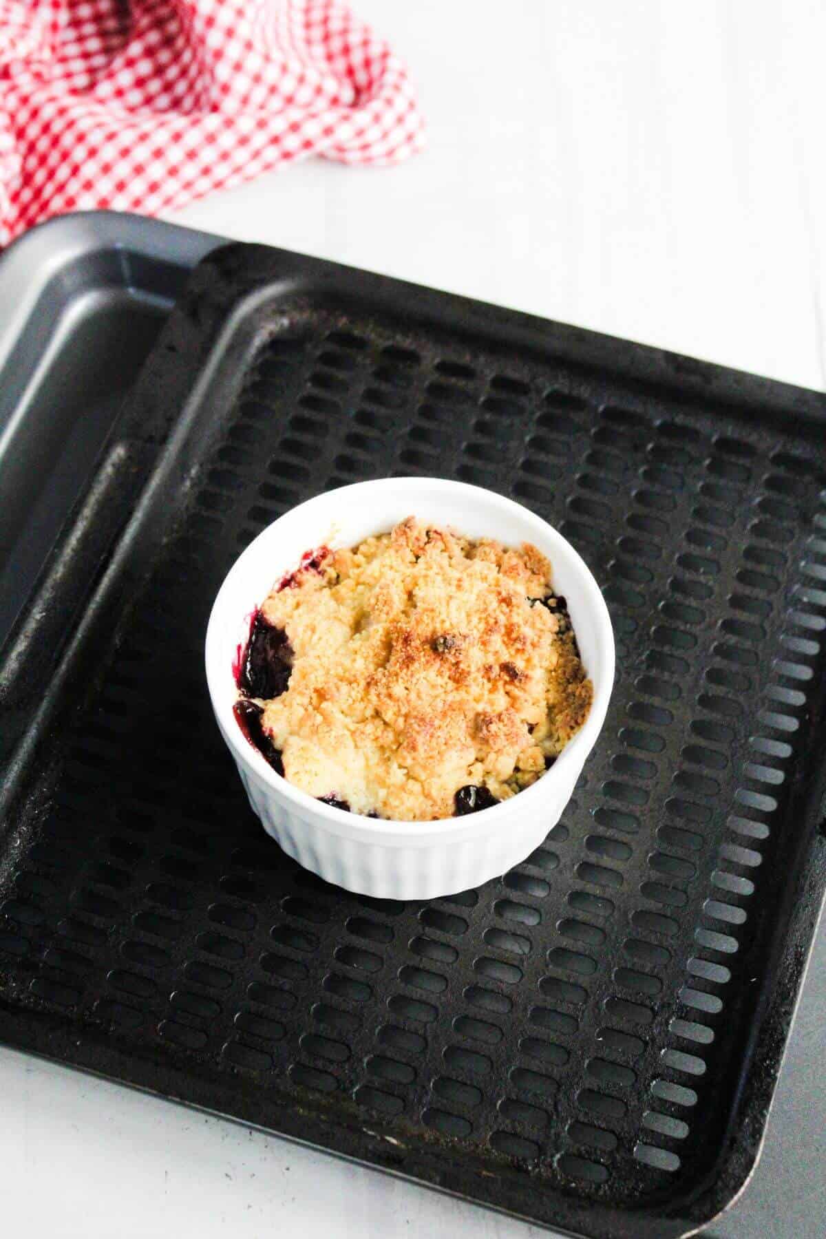 A single-serving berry crumble in a white ramekin sits on a black baking tray with a red-and-white cloth in the background.