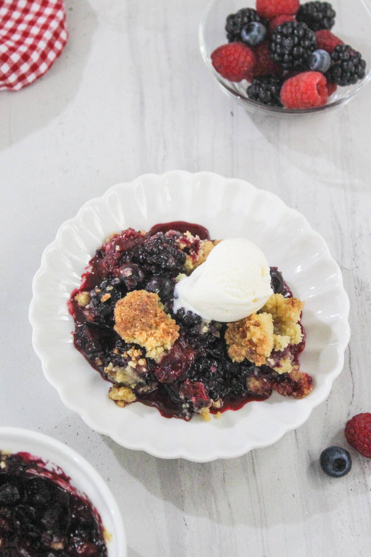 A serving of berry cobbler topped with a scoop of vanilla ice cream is displayed in a white bowl, with fresh berries in a glass bowl nearby on a light surface.