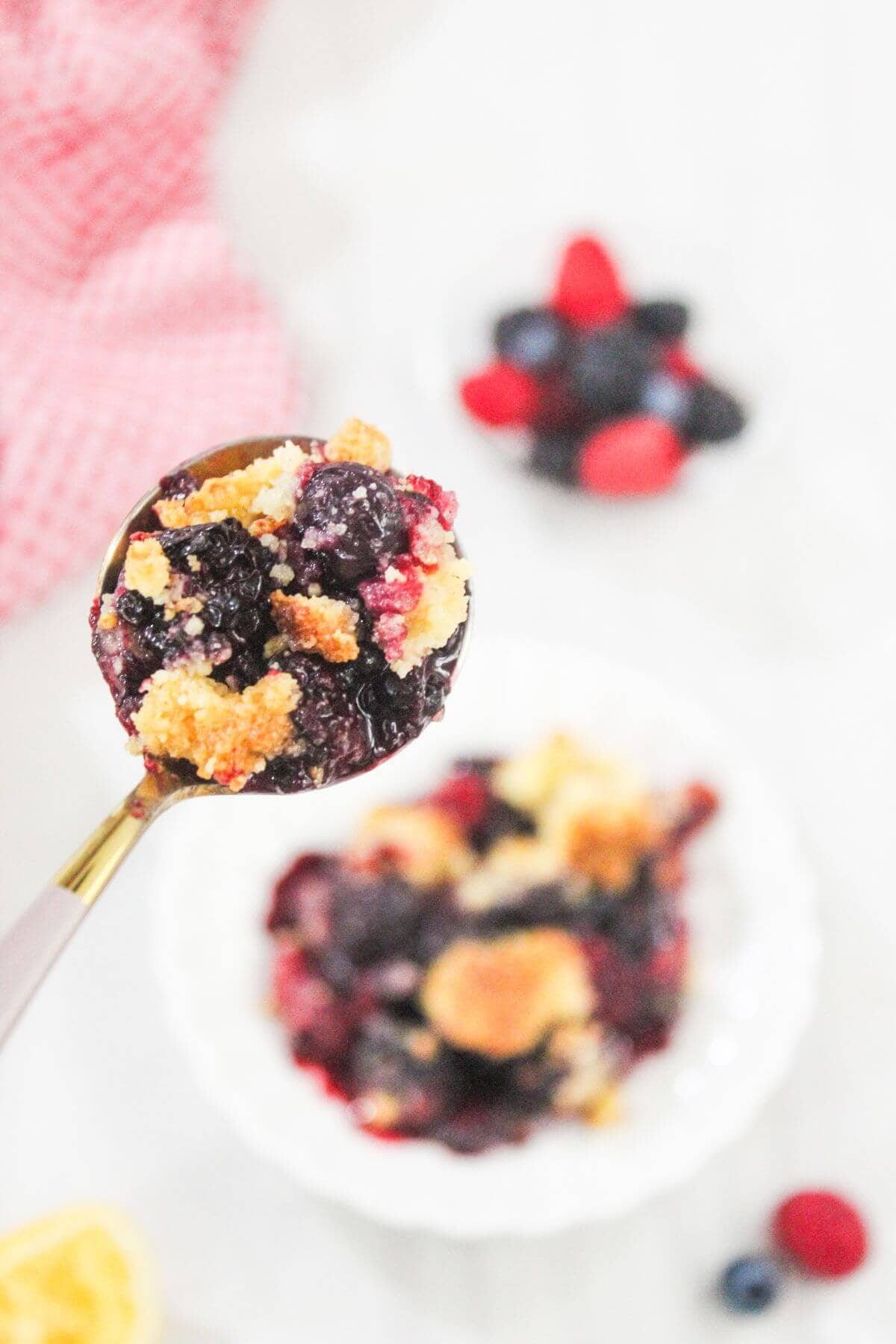 A spoonful of mixed berry crumble held above a bowl with more crumble, fresh berries, and a red cloth in the background.