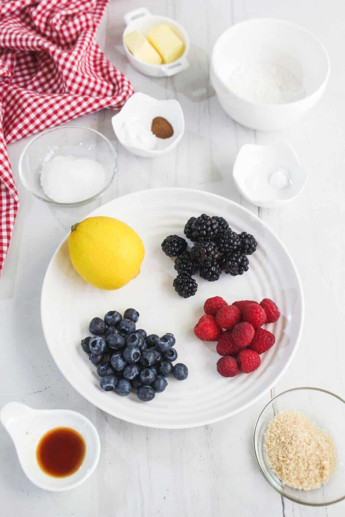 A plate with blueberries, blackberries, raspberries, and a lemon, surrounded by small bowls of sugar, butter, flour, brown sugar, vanilla, and spices on a white surface.