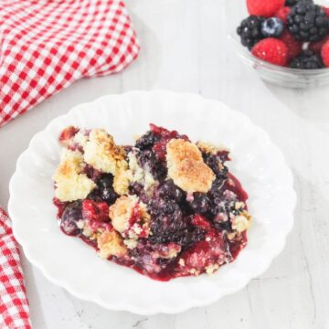 A white plate with berry cobbler sits on a white surface next to a red checkered cloth; a bowl of mixed berries is in the background.
