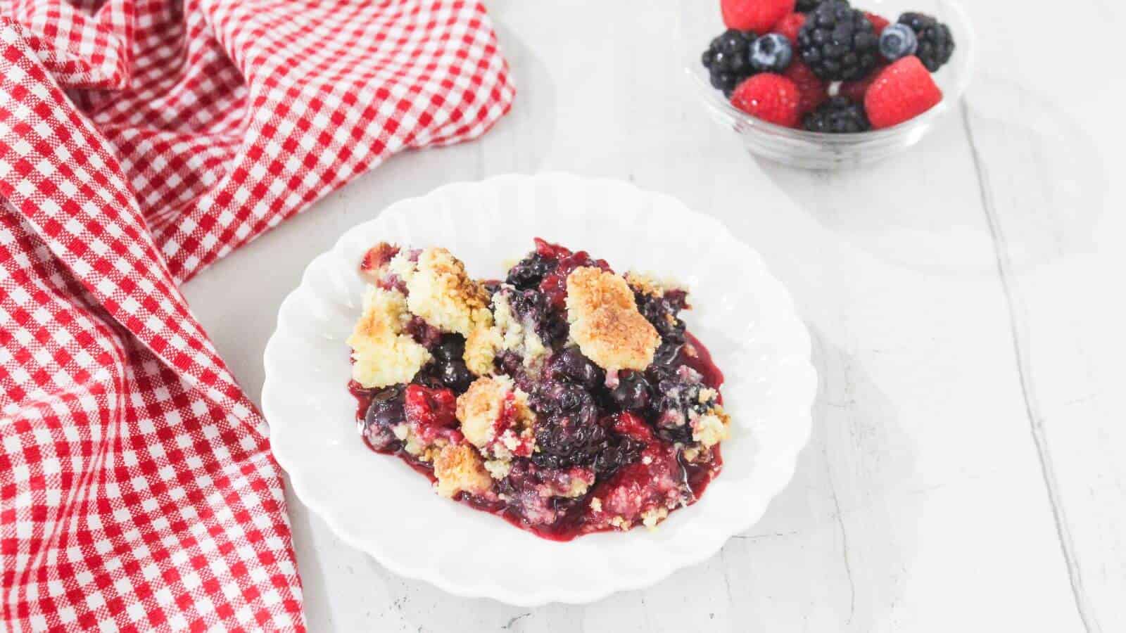 A white plate with berry cobbler sits on a white surface next to a red checkered cloth; a bowl of mixed berries is in the background.