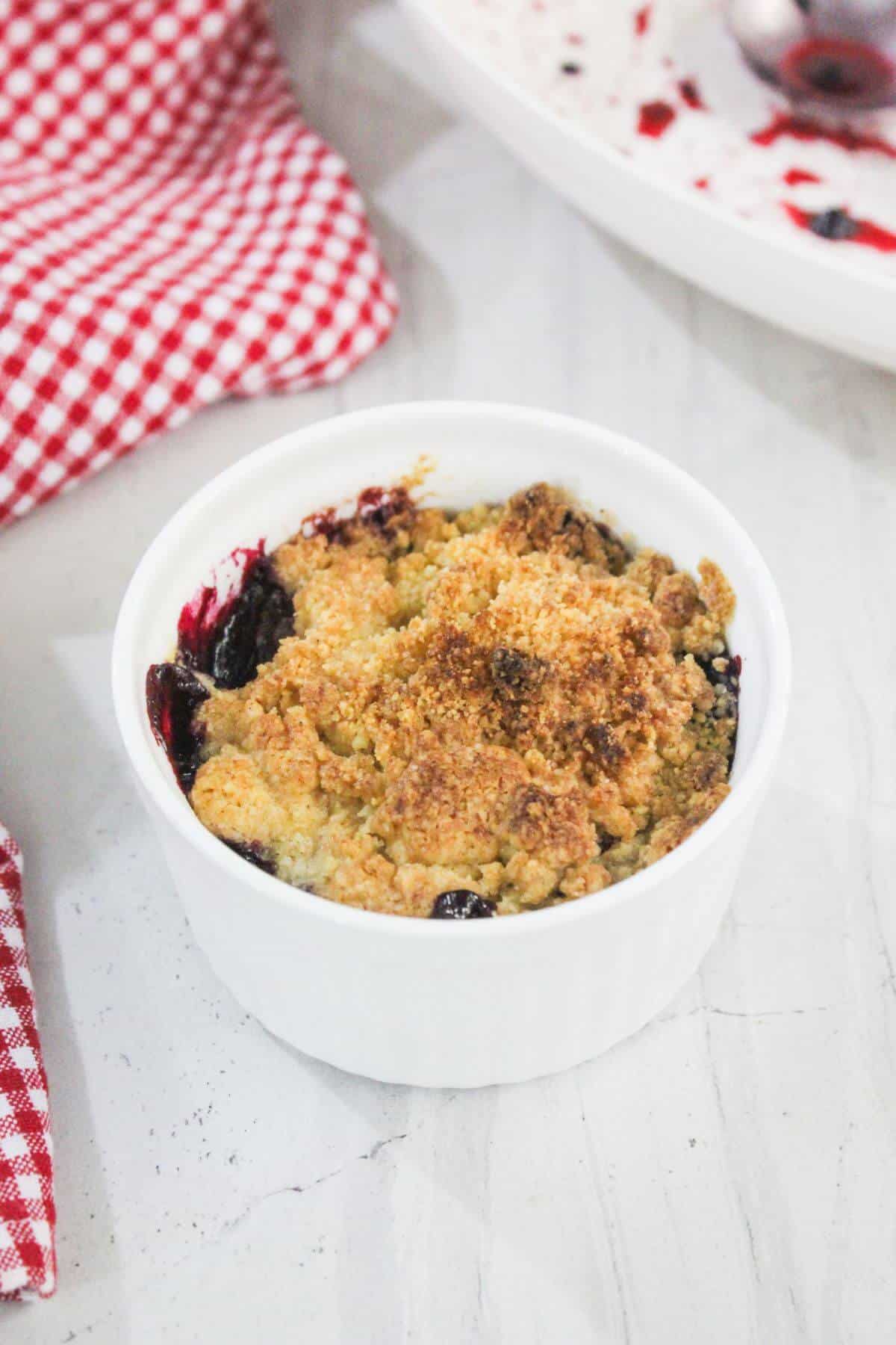 A small white ramekin filled with baked berry crumble sits on a white surface next to a red and white checkered cloth.