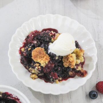 A serving of berry cobbler topped with a scoop of vanilla ice cream is presented on a white scalloped plate.
