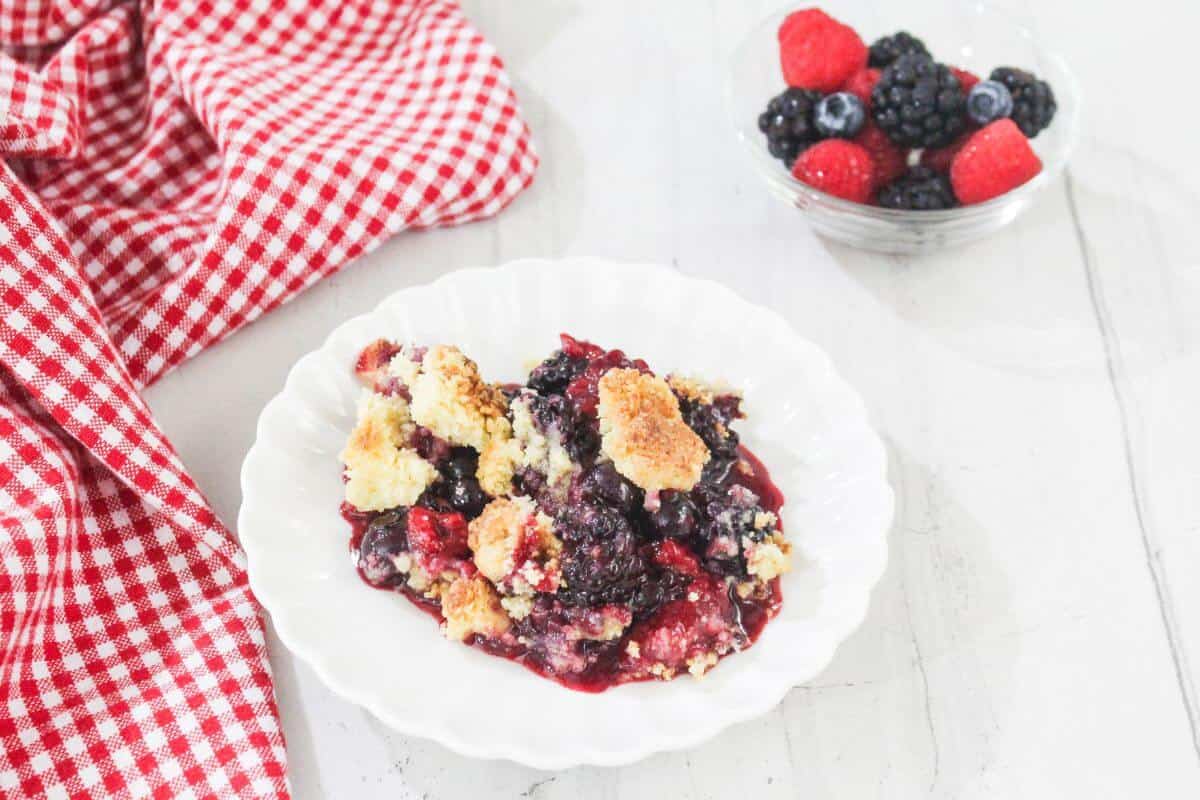 A serving of berry crumble on a white plate sits next to a bowl of fresh berries and a red checkered cloth on a white surface.