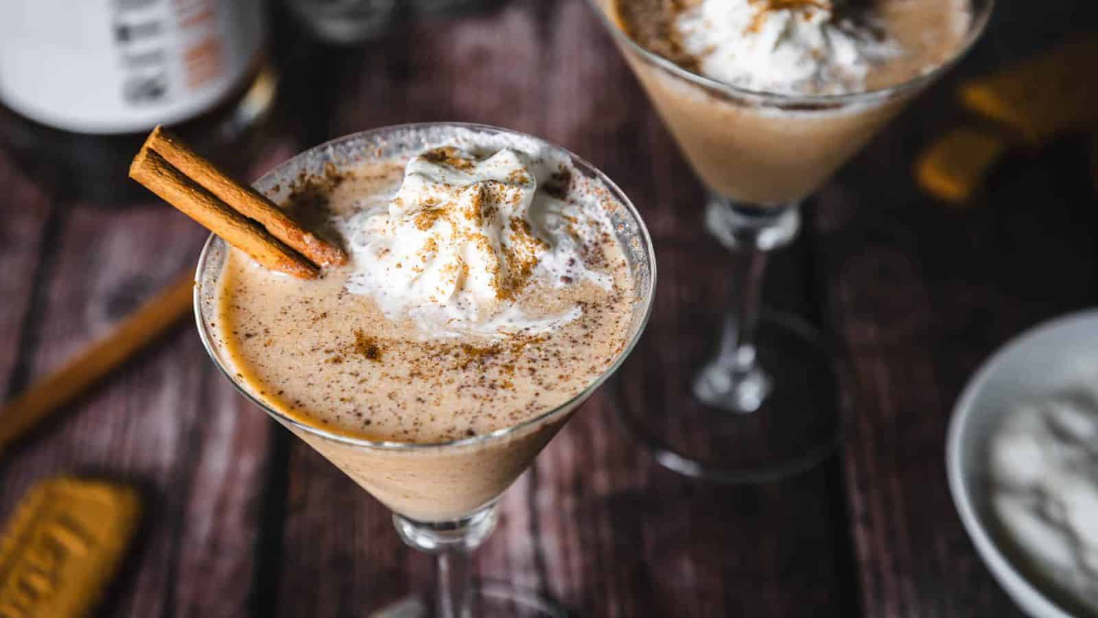 A creamy brown cocktail in a martini glass topped with whipped cream, a sprinkle of cinnamon, and a cinnamon stick, set on a wooden surface.