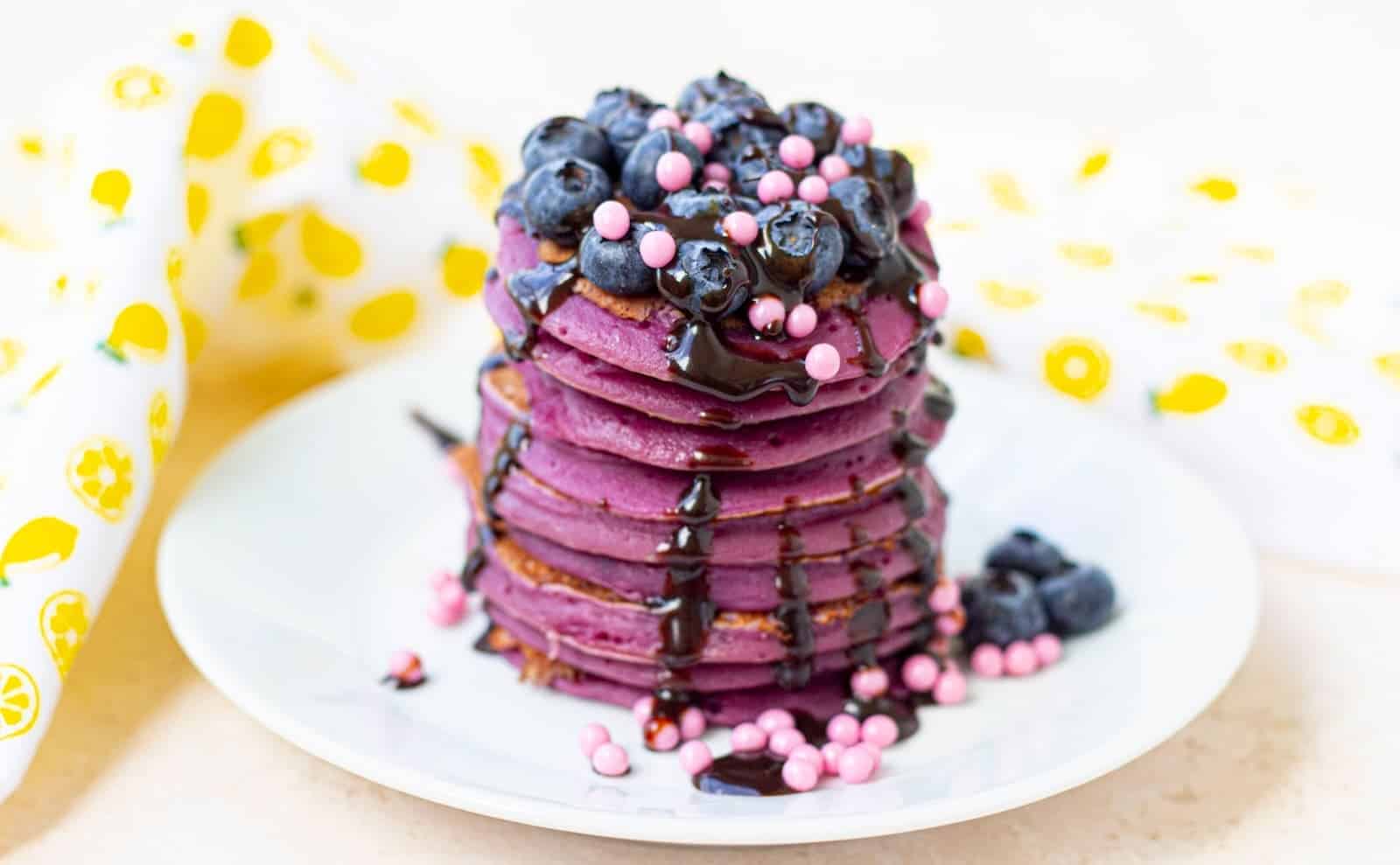A stack of purple pancakes topped with blueberries, pink candy pearls, and chocolate syrup on a white plate.