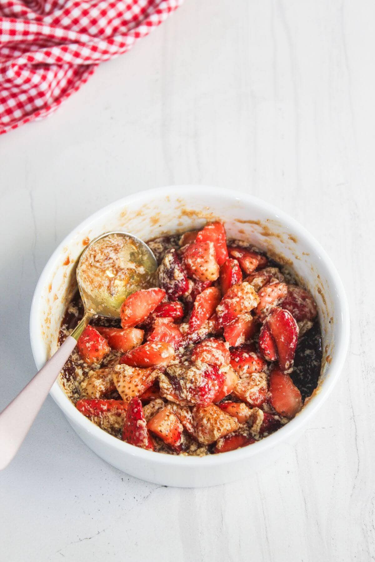 A white bowl filled with sliced strawberries, sweetener, and crushed nuts, with a metal spoon resting inside. A red checkered cloth is partially visible in the background.