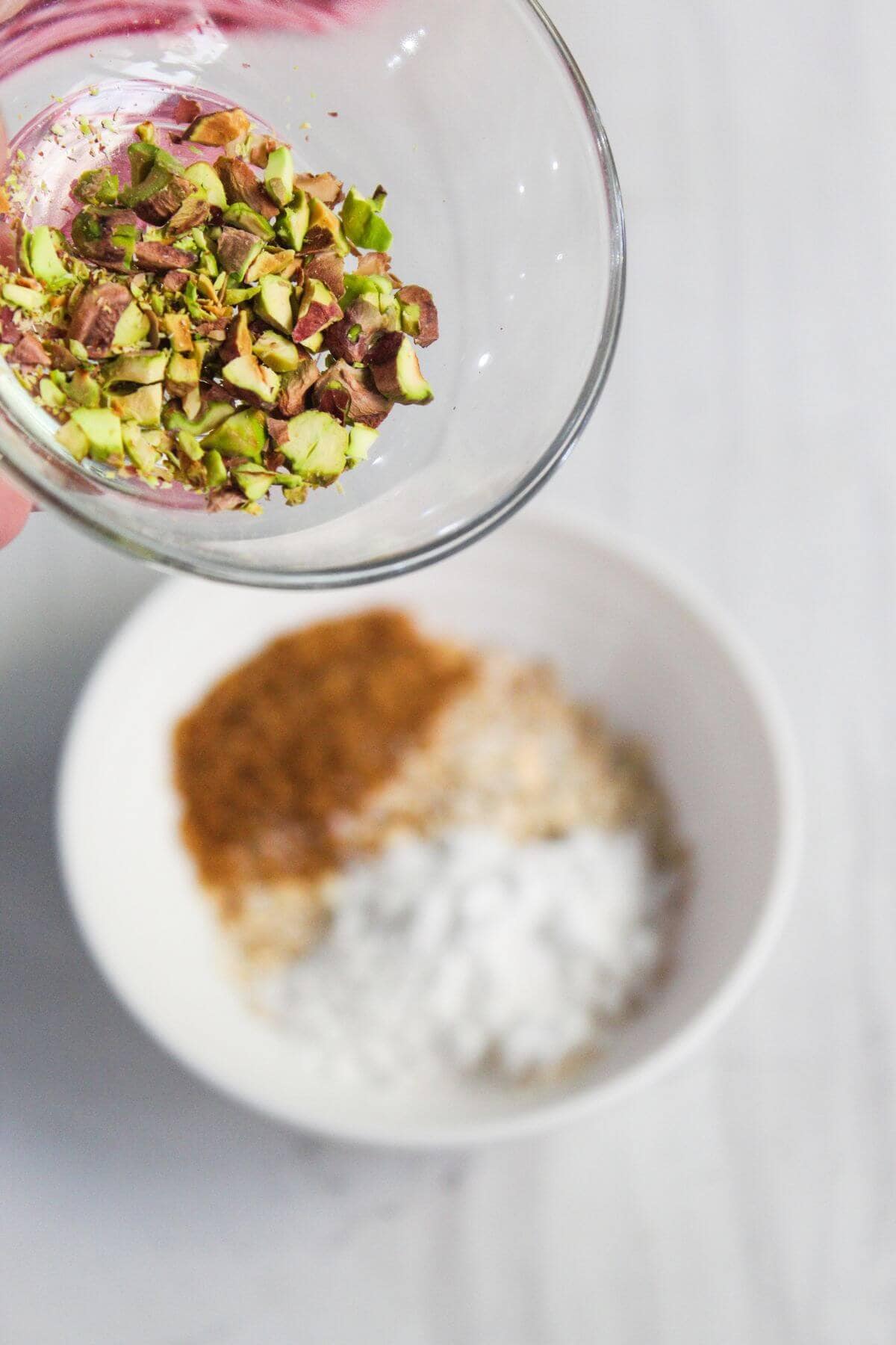 A hand holding a glass bowl of chopped pistachios above a white bowl containing brown sugar, white sugar, and flour.