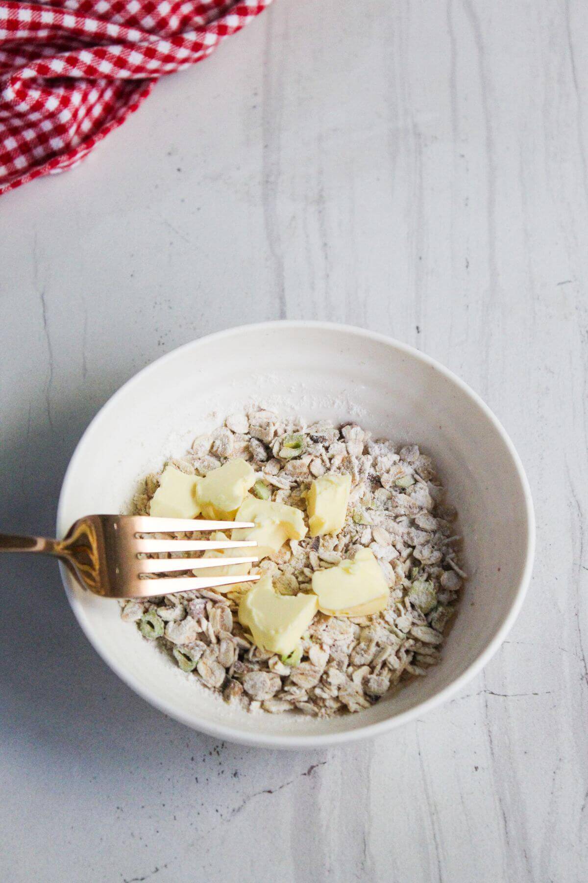 A white bowl with oats, flour, seeds, and cubes of butter being mixed with a gold fork on a light countertop.