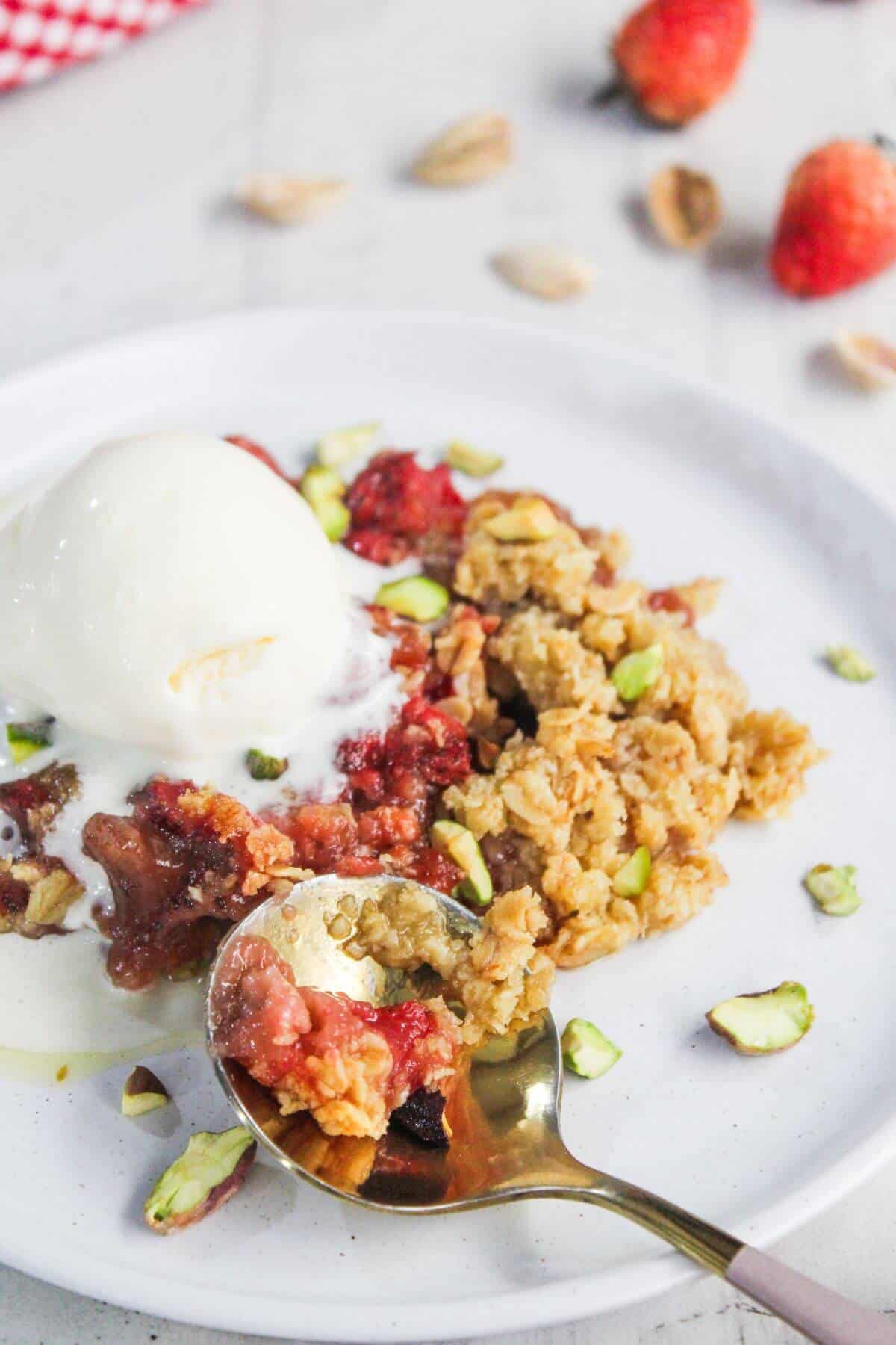 A plate with fruit crumble topped with chopped pistachios and served with a scoop of vanilla ice cream; a spoonful is being lifted from the plate.