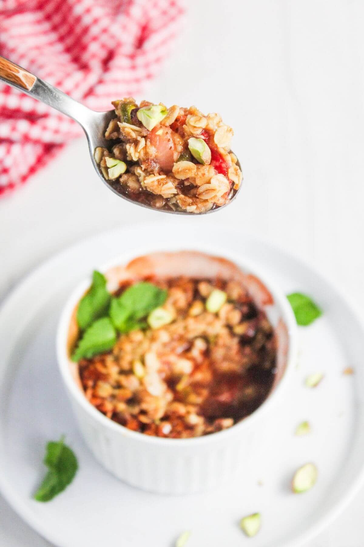 A spoonful of fruit and oat crumble is held above a small white ramekin filled with the same dessert, garnished with green herbs and chopped nuts.