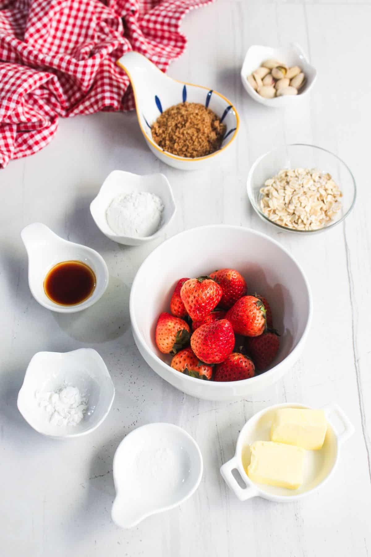 Assorted baking ingredients for strawberry crisp, including fresh strawberries, oats, brown sugar, butter, vanilla extract, pistachios, and small bowls of powders on a white surface with a red checked cloth.