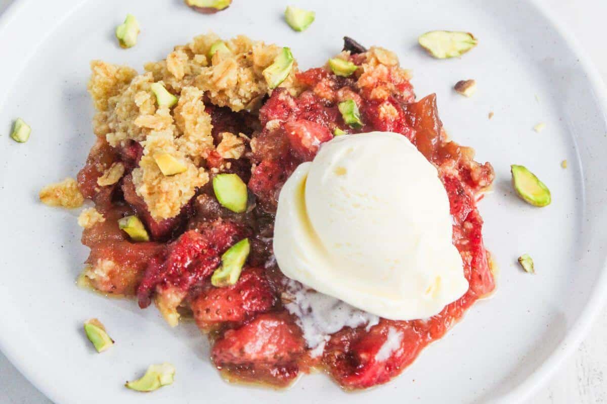 A serving of fruit crumble topped with a scoop of vanilla ice cream, garnished with chopped pistachios, on a white plate.