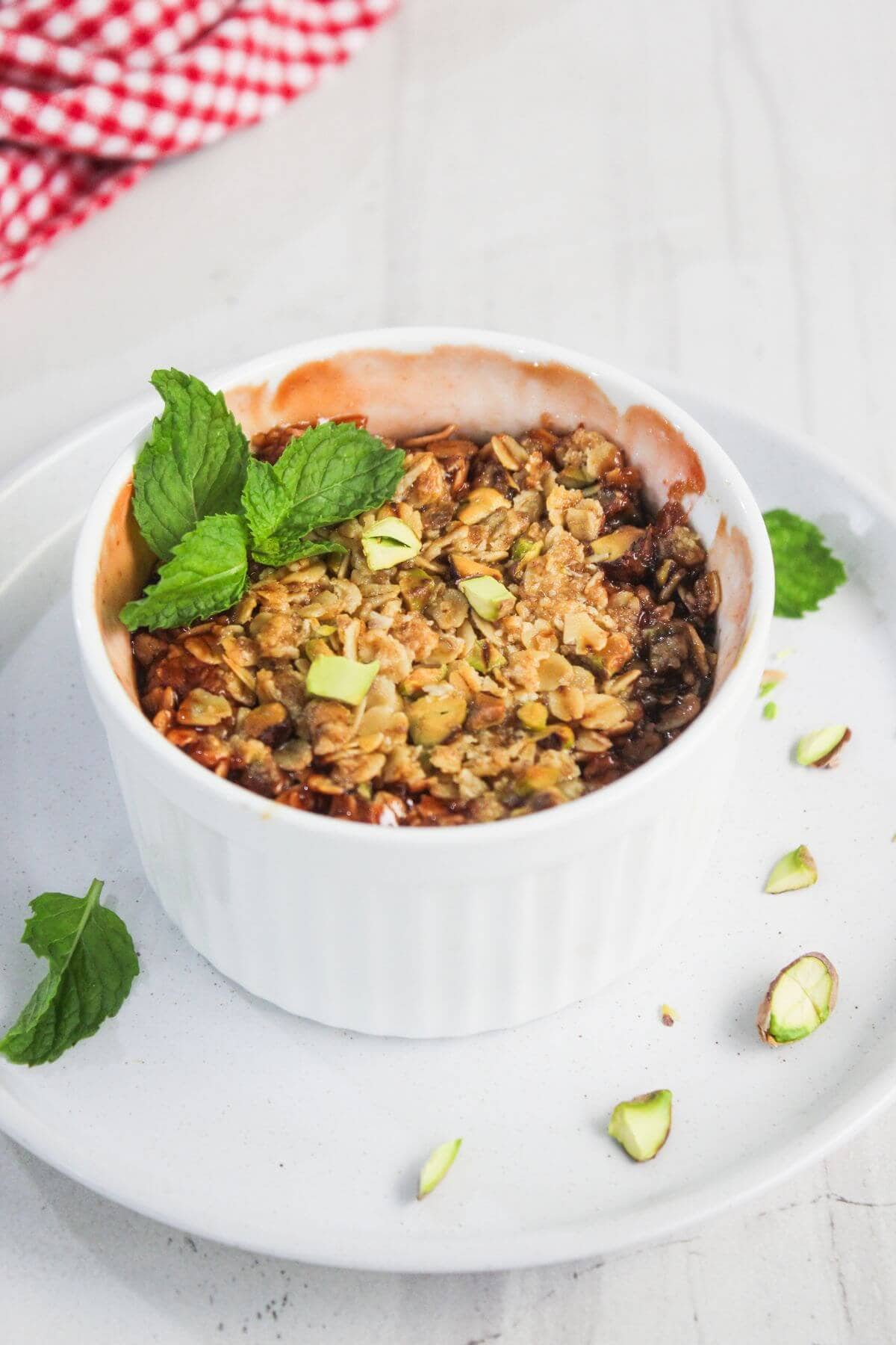 A baked fruit crumble topped with oats, pistachios, and mint leaves, served in a white ramekin on a white plate.