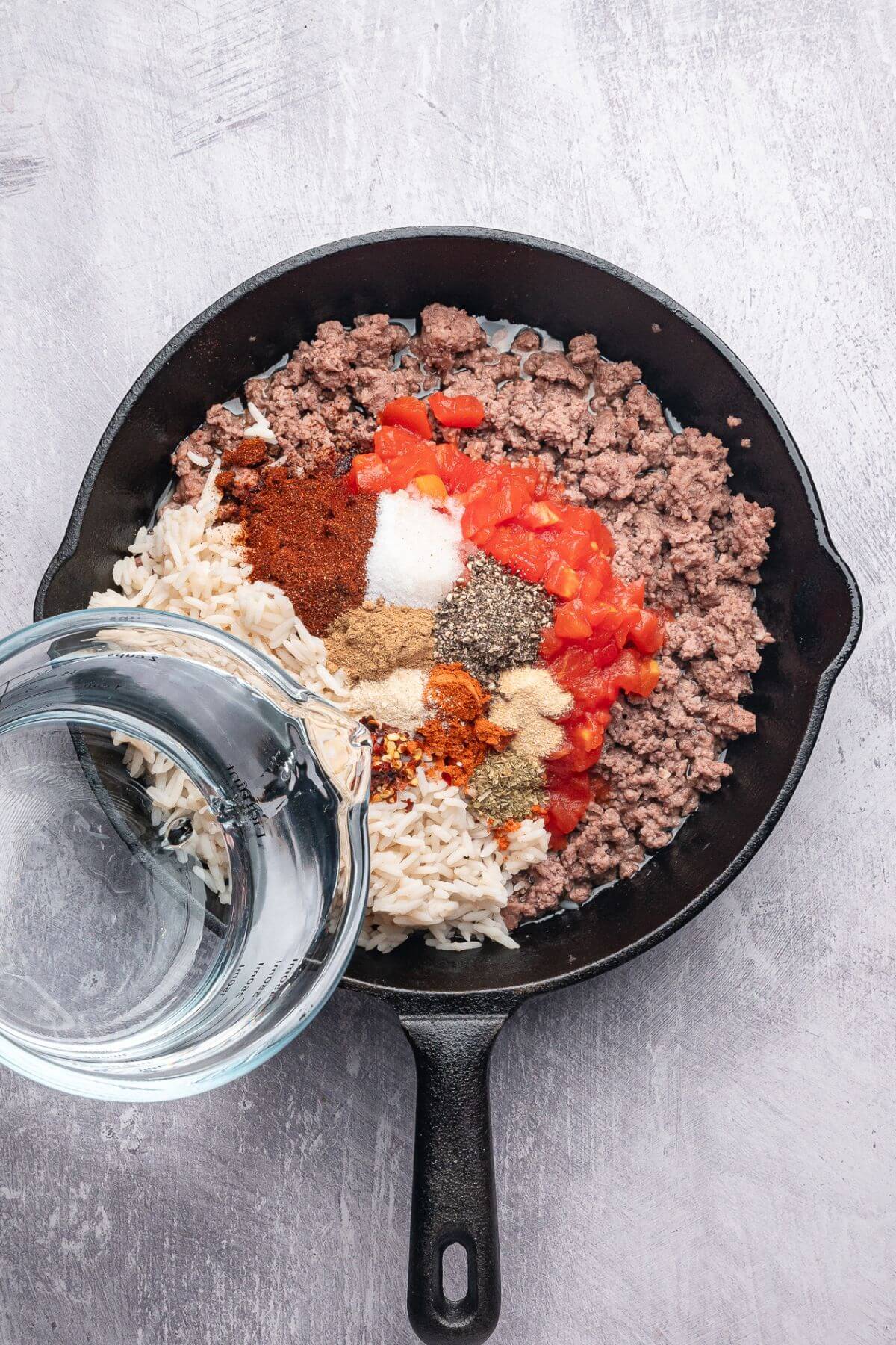 A cast iron skillet with cooked ground beef, white rice, diced tomatoes, assorted spices, and water being poured in.