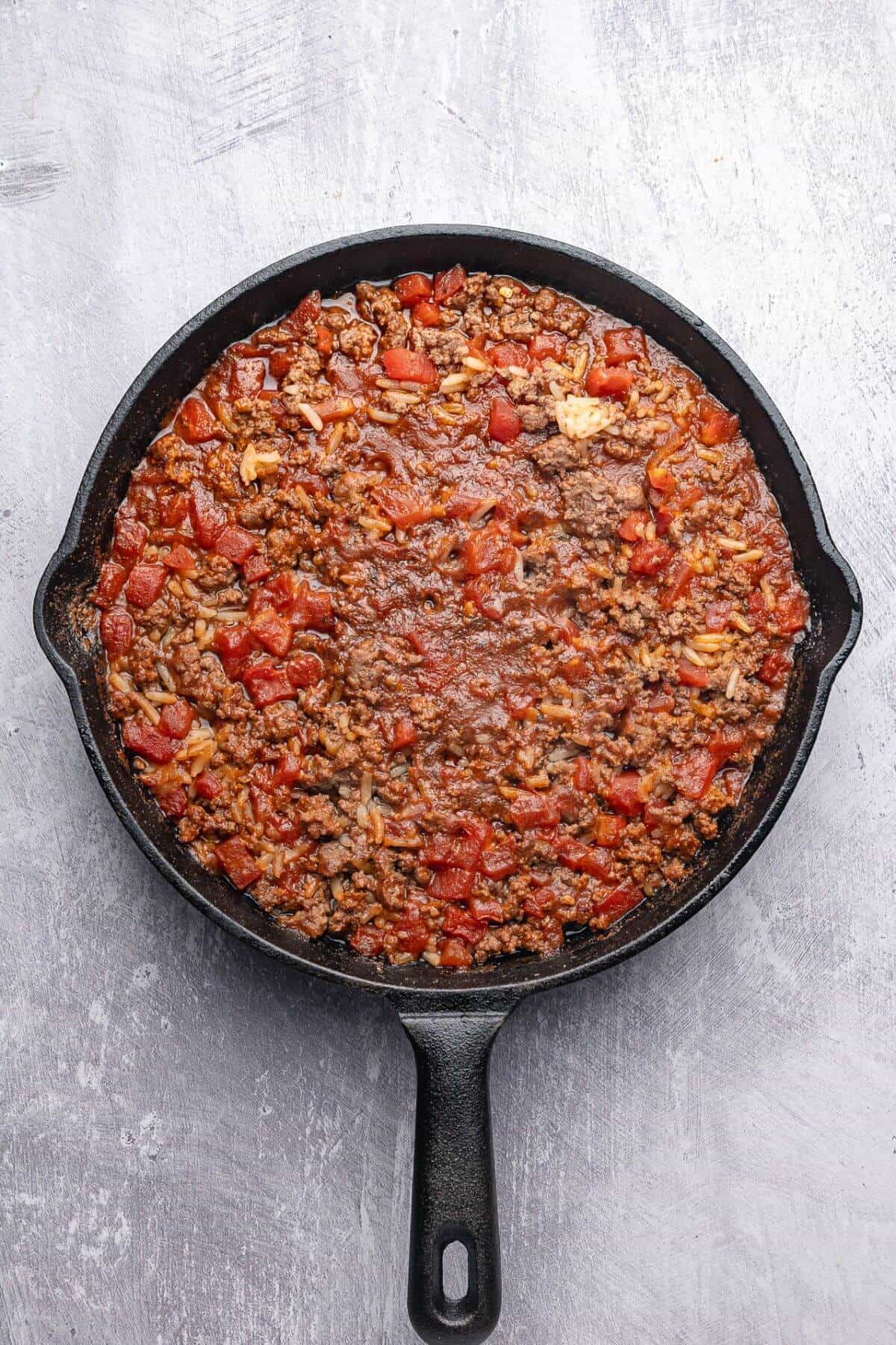A cast iron skillet filled with cooked ground beef, diced tomatoes, and seasonings on a gray textured surface.
