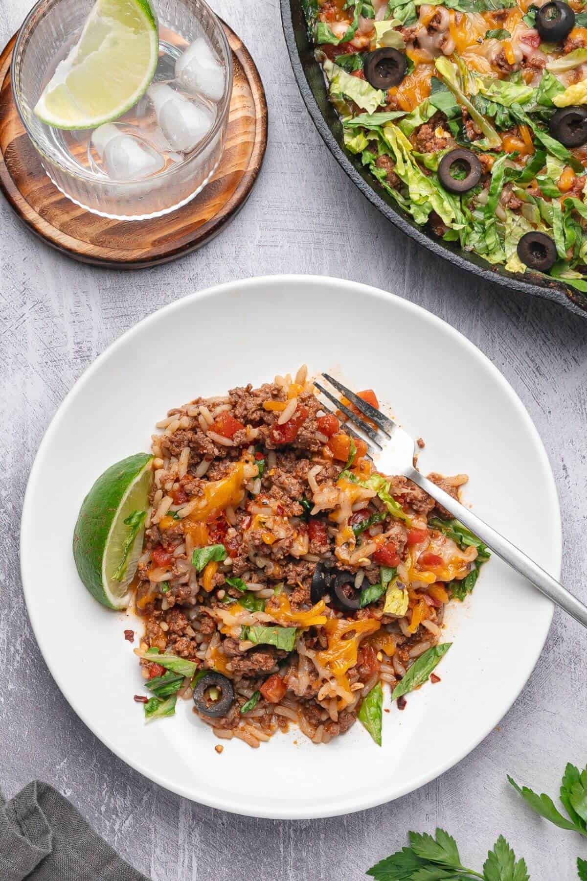A plate of taco rice topped with ground beef, cheese, lettuce, and black olives, served with a lime wedge. A glass of water with ice and lime and a skillet of taco rice are nearby.