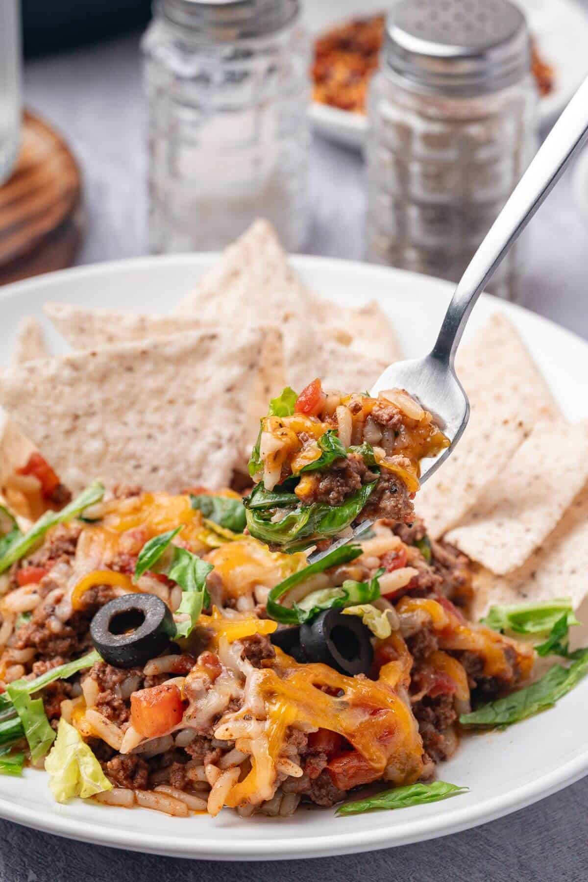 A fork holds a bite of taco salad with ground beef, cheese, lettuce, diced tomatoes, olives, and tortilla chips on a white plate.