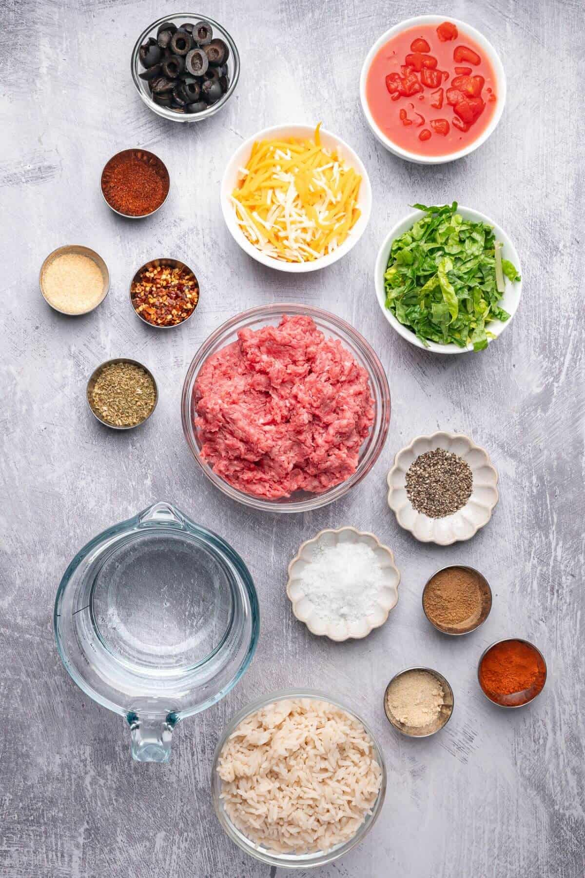 Various ingredients including ground meat, shredded cheese, rice, chopped lettuce, sliced olives, tomatoes, spices, and water arranged on a gray surface.