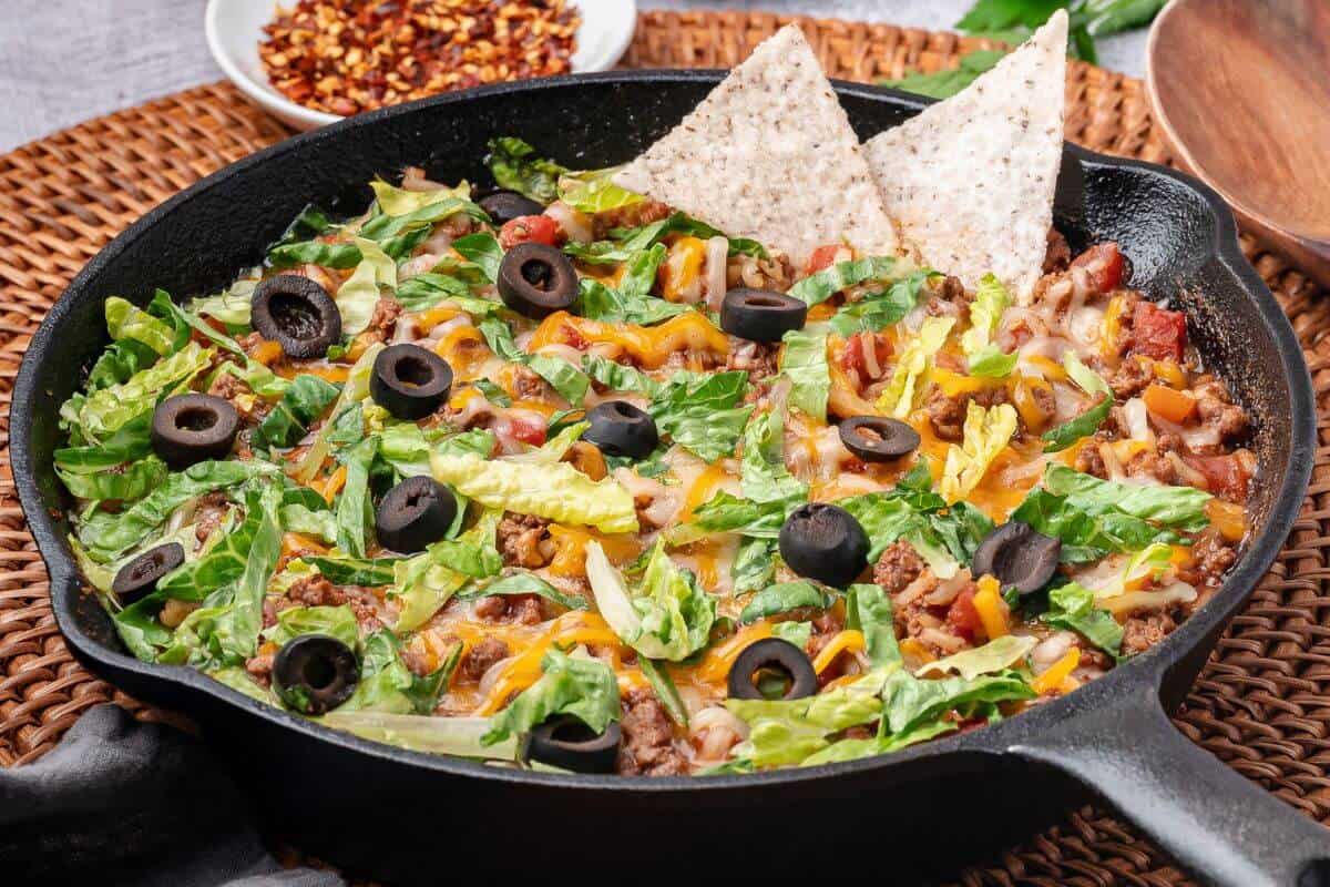 A cast iron skillet filled with taco salad topped with lettuce, shredded cheese, black olives, and two tortilla chips, placed on a woven mat.