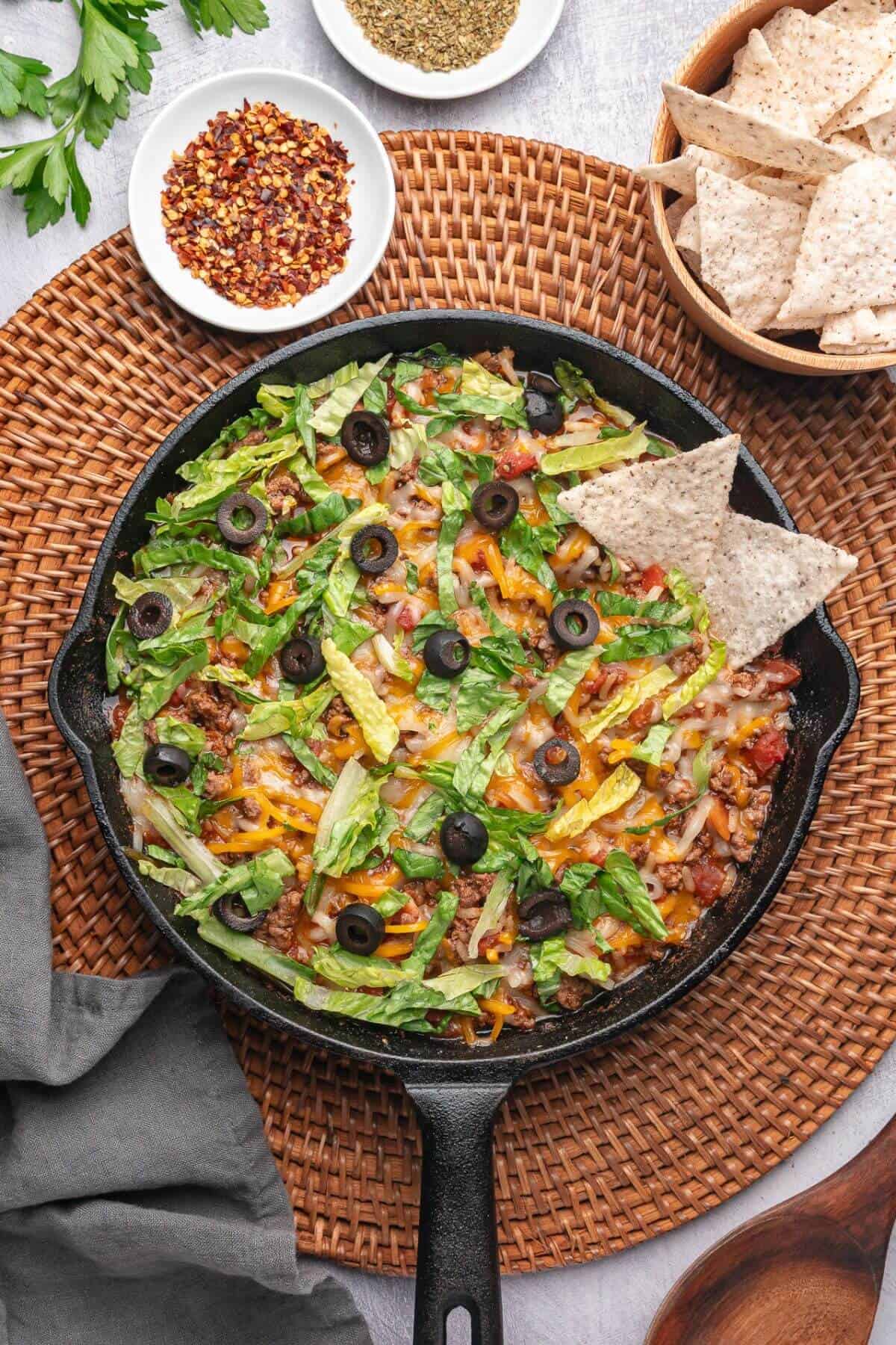 A skillet filled with taco salad topped with lettuce, cheese, olives, and tortilla chips, placed on a woven mat with bowls of red pepper flakes, oregano, and extra chips nearby.