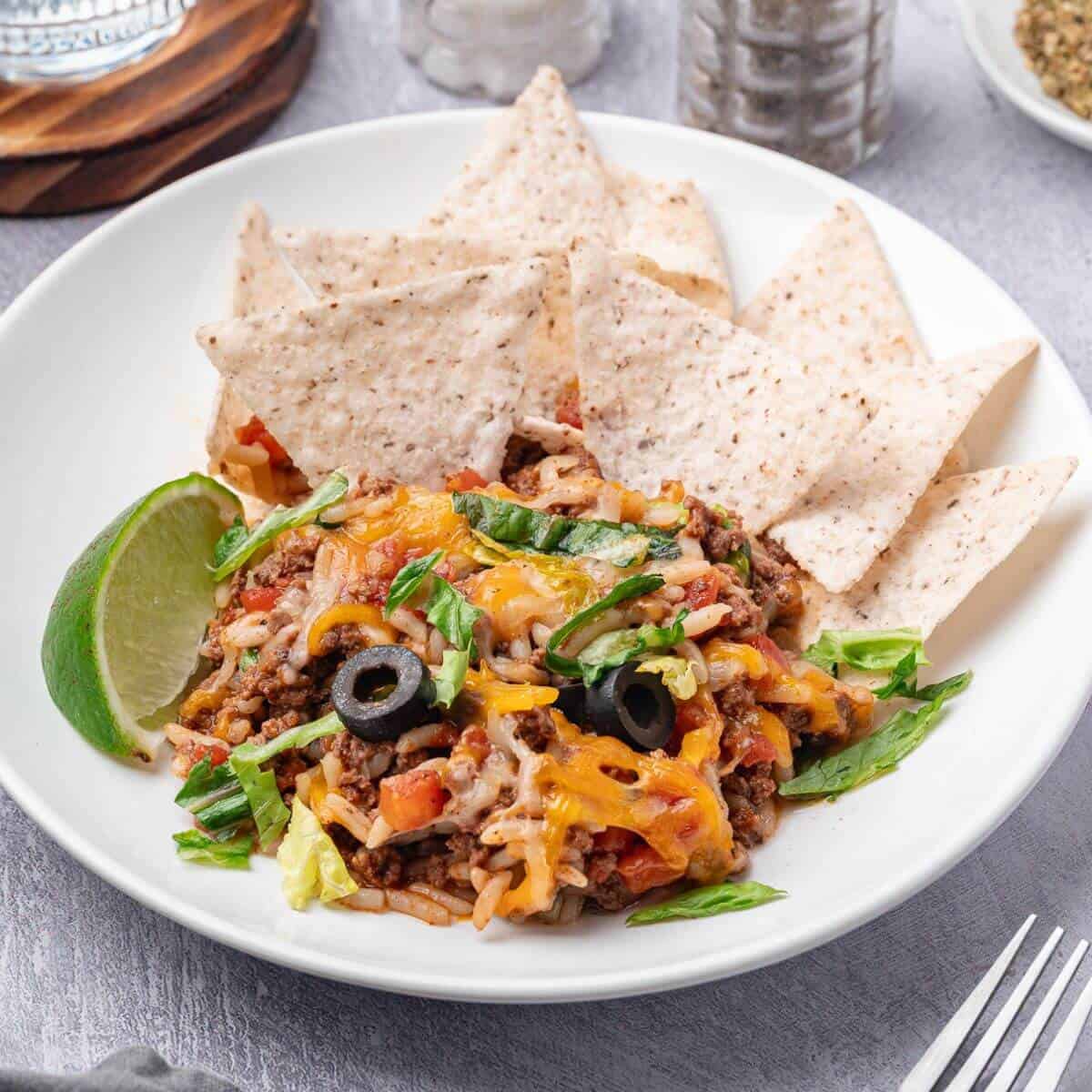 A plate of taco casserole topped with cheese, black olives, and green onions, served with tortilla chips and a lime wedge.