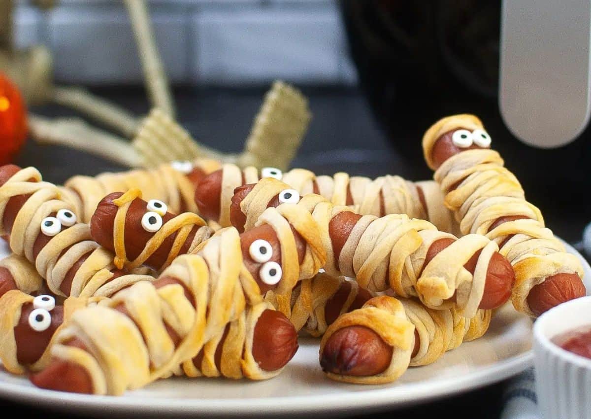 A plate of hot dogs wrapped in pastry to resemble mummies, with candy eyes on each, served as a Halloween-themed snack.