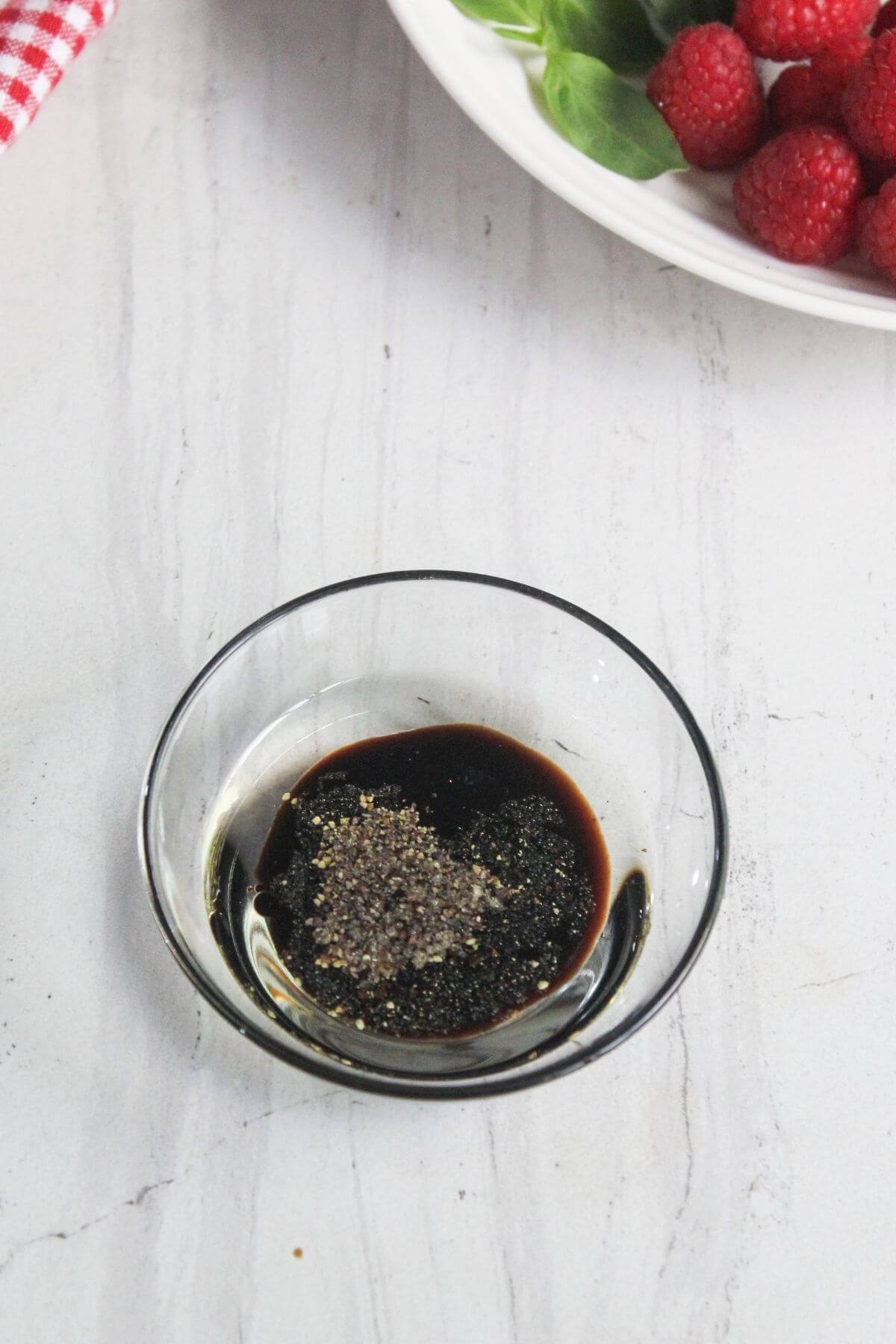 A small glass bowl contains a mixture of dark liquid and cracked black pepper on a light countertop, with a plate of fresh raspberries and greens partly visible in the background.