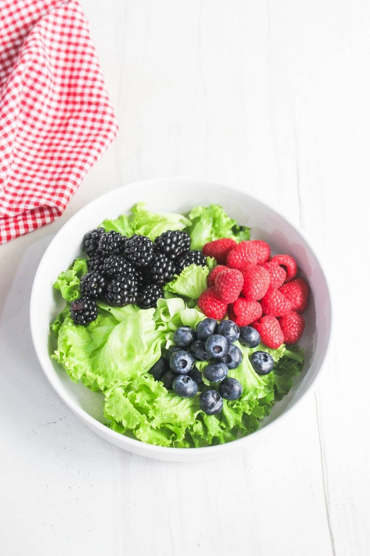 A white bowl filled with green leaf lettuce, blackberries, raspberries, and blueberries sits on a white surface next to a red and white checked cloth.