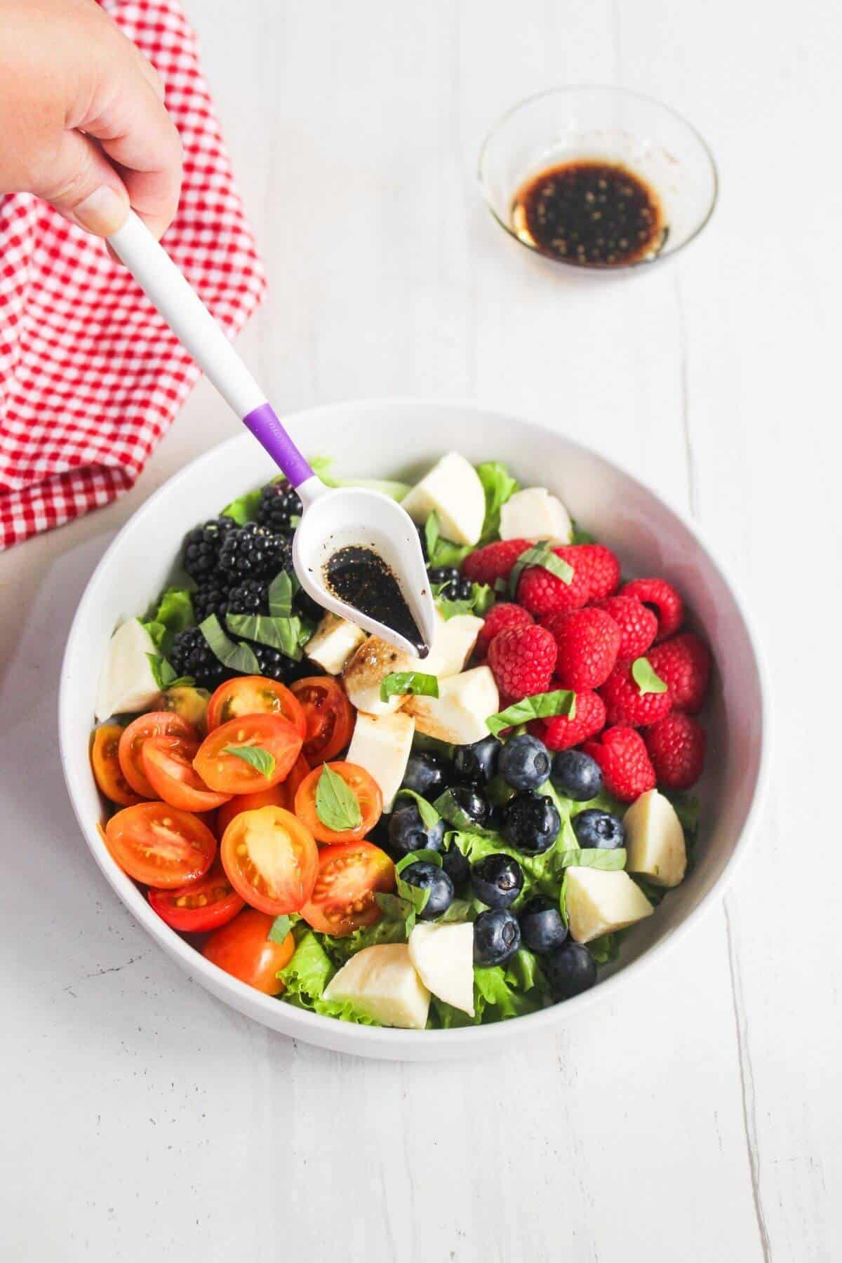 A bowl of salad with tomatoes, mozzarella, blackberries, blueberries, raspberries, and basil, with dressing being poured on top; a bowl of dressing and a red cloth are nearby.