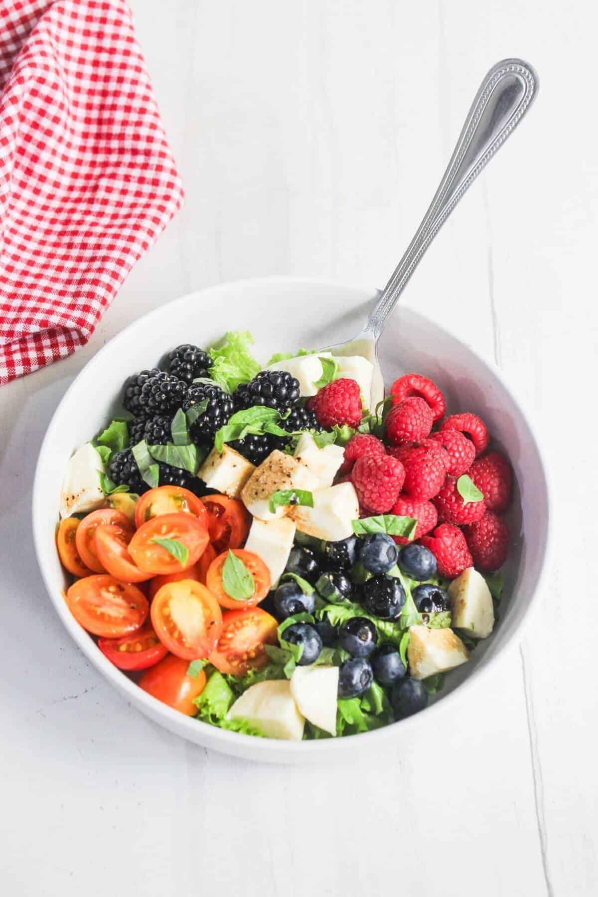 A white bowl filled with cherry tomatoes, blackberries, blueberries, raspberries, mozzarella chunks, and leafy greens with a fork, set on a white surface next to a red checkered cloth.