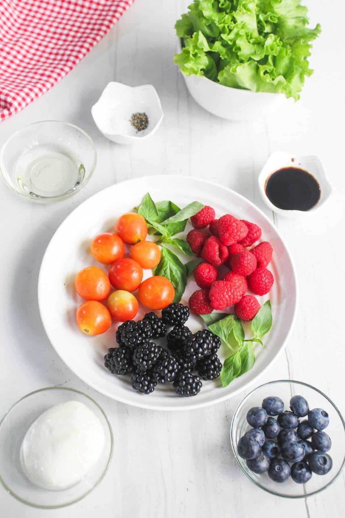 A white plate with cherry tomatoes, raspberries, blackberries, blueberries, and basil, surrounded by bowls of lettuce, mozzarella, vinegar, oil, pepper, and salt on a white table.