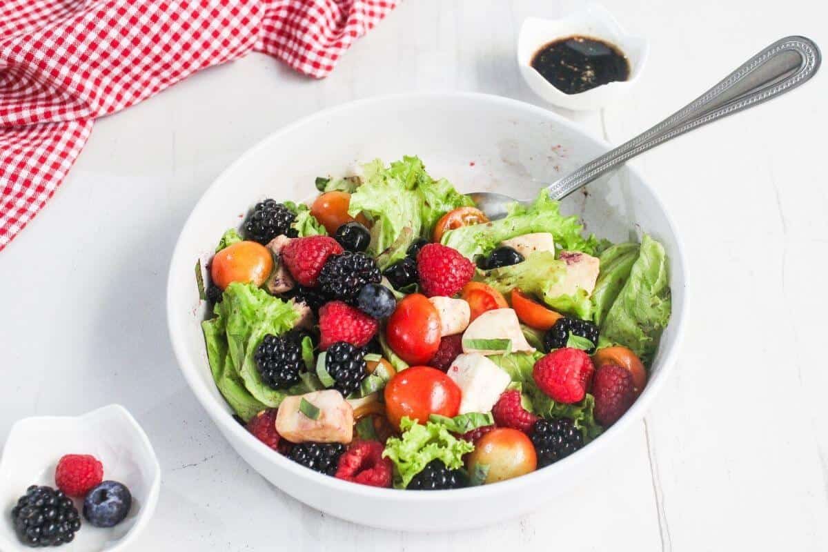 A white bowl filled with leafy salad, cherry tomatoes, berries, avocado, and a metal spoon, with a small dish of dark dressing and a red checkered cloth nearby.