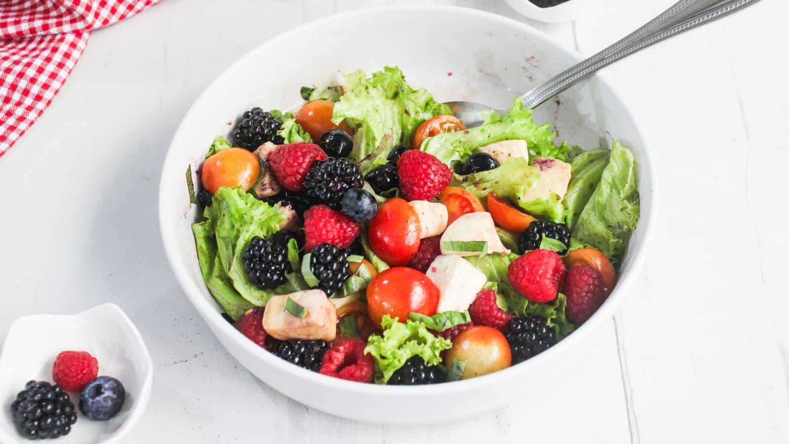 A white bowl filled with leafy salad, cherry tomatoes, berries, avocado, and a metal spoon, with a small dish of dark dressing and a red checkered cloth nearby.