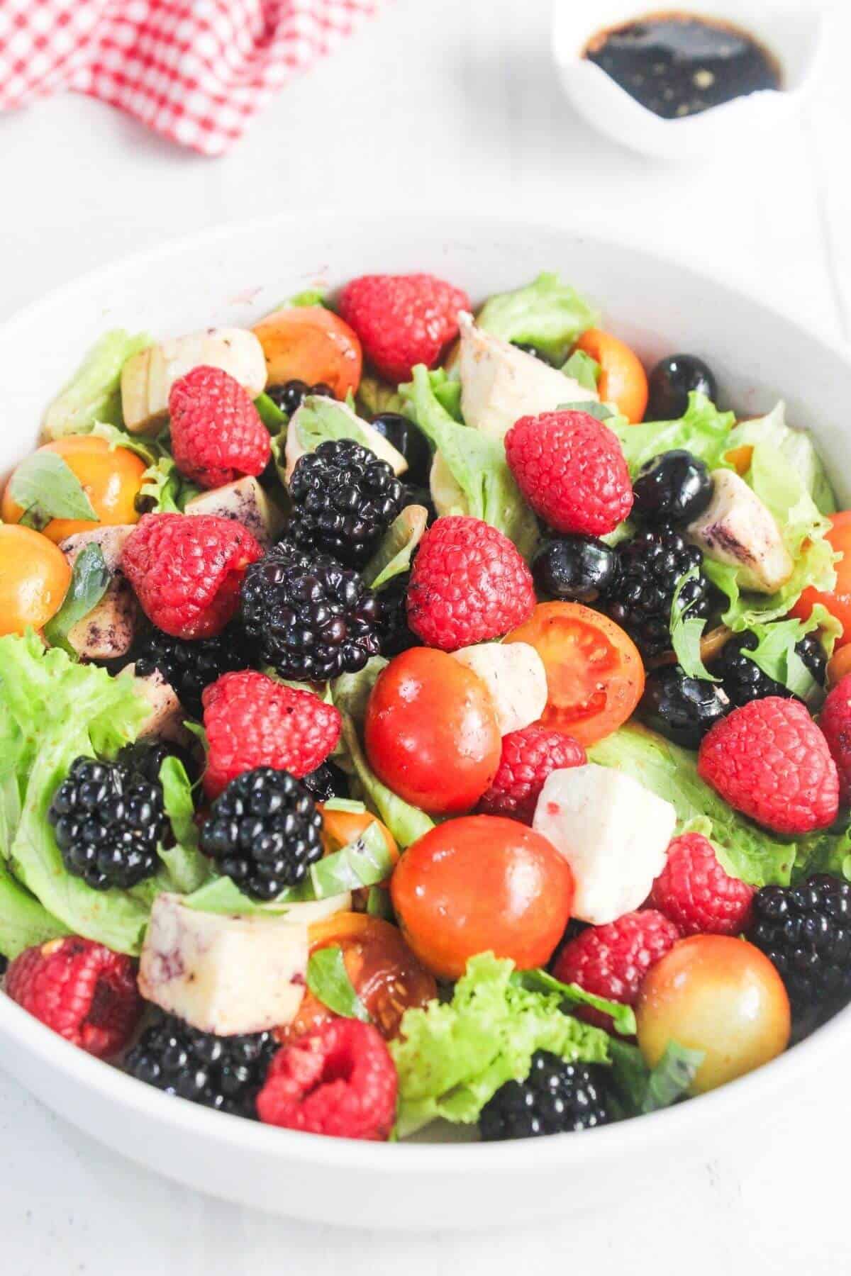 A bowl of salad with lettuce, cherry tomatoes, blackberries, raspberries, blueberries, and mozzarella cheese, with a small dish of dark dressing in the background.
