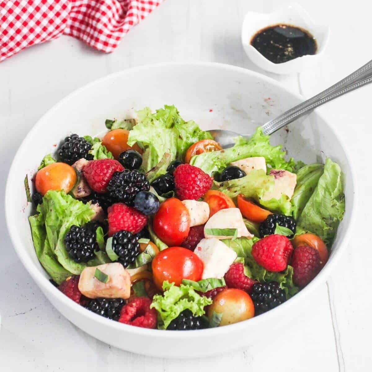 A bowl of mixed green salad with cherry tomatoes, blackberries, raspberries, blueberries, mozzarella chunks, and basil, with a small dish of dark dressing on the side.