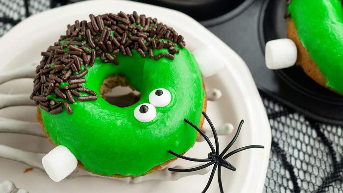 A donut with green icing, chocolate sprinkles, candy eyes, marshmallow "ears," and a plastic spider on a white plate.