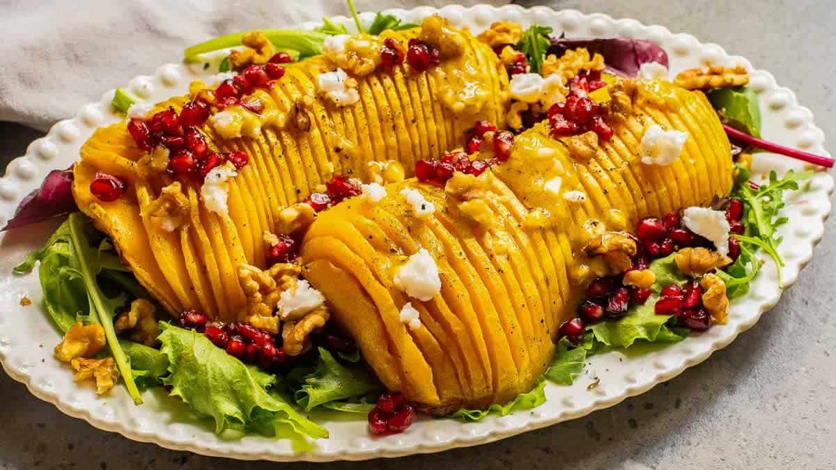 Sliced roasted squash on mixed greens, topped with crumbled cheese, pomegranate seeds, and walnuts, served on a white platter.