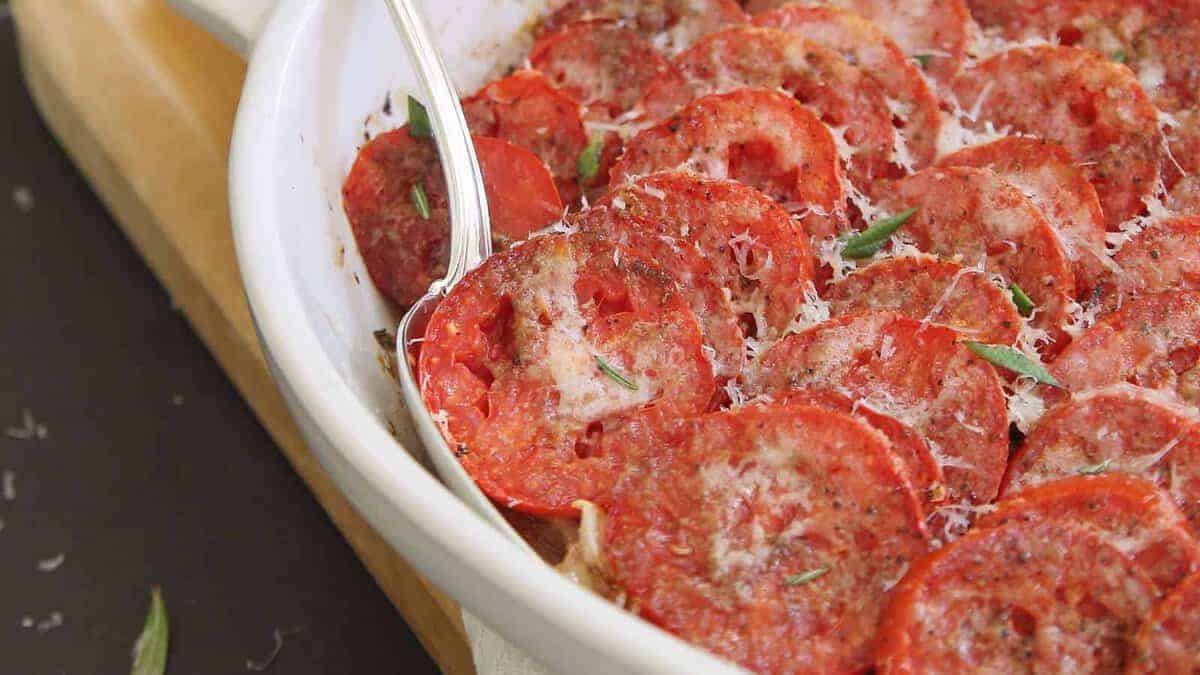 A white baking dish filled with sliced tomatoes topped with grated cheese and herbs, with a serving spoon on the side.