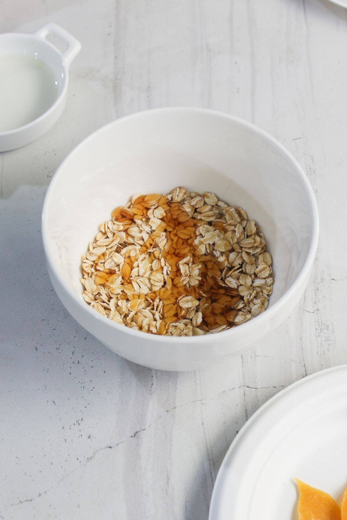 A white bowl containing rolled oats and honey sits on a light-colored marble surface, with a small dish of milk nearby.